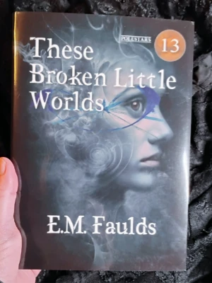 Cover of the hardback special edition of These Broken Little Worlds by E.M. Faulds. It is held in a hand over a shiny black lace backdrop. Polestars 13 is written in the top right corner. The cover image is of a woman's face in profile with cracks and swirls over it.